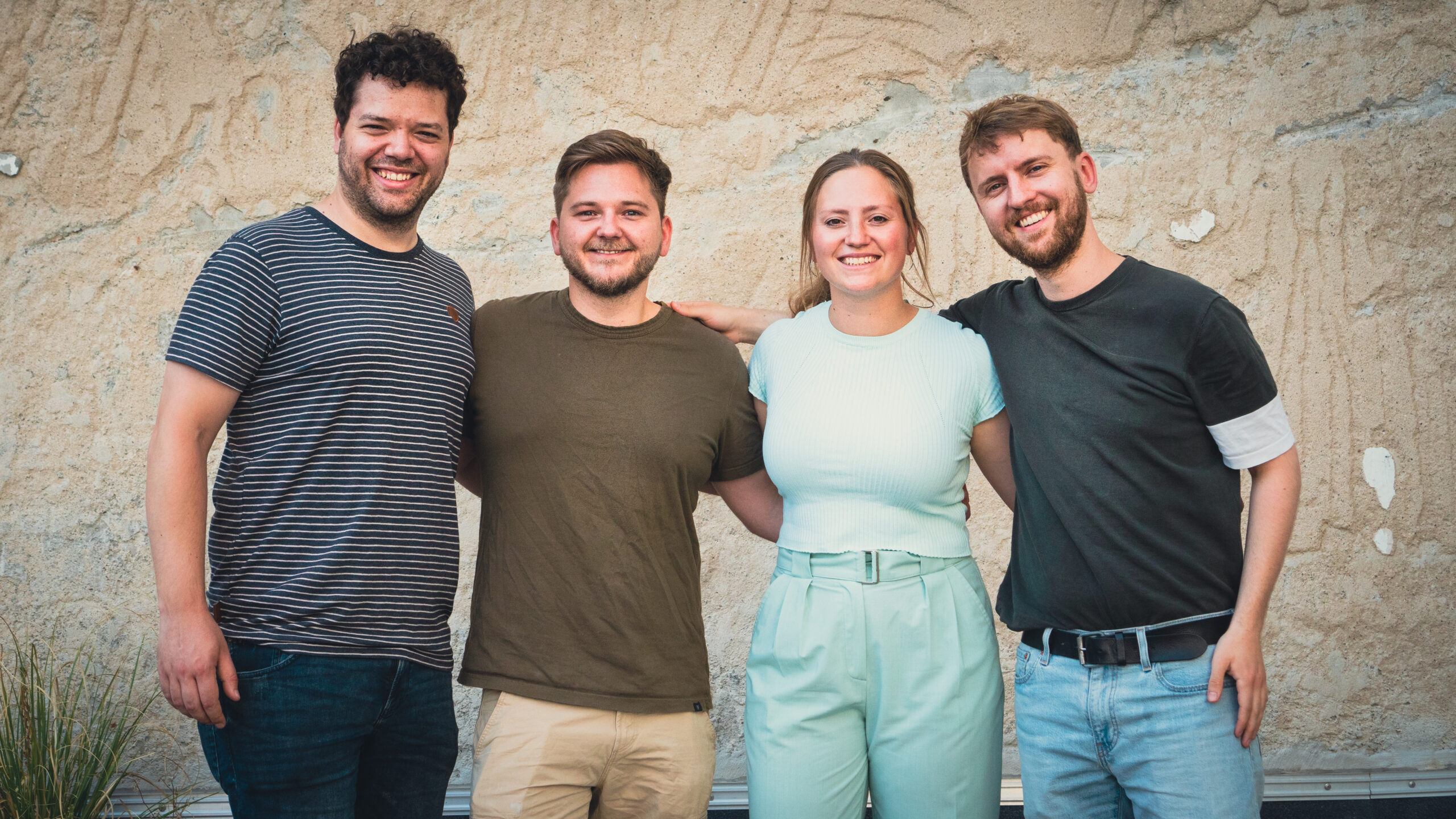 Teamfoto holoNative mit Menschen vor einer Wand, die sich umarmen und grinsen.